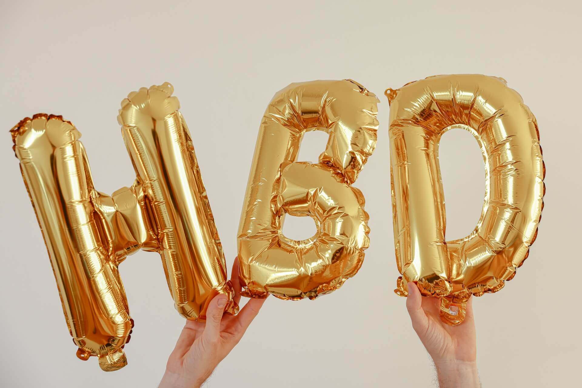 Two hands hold up gold balloons spelling “HBD”.